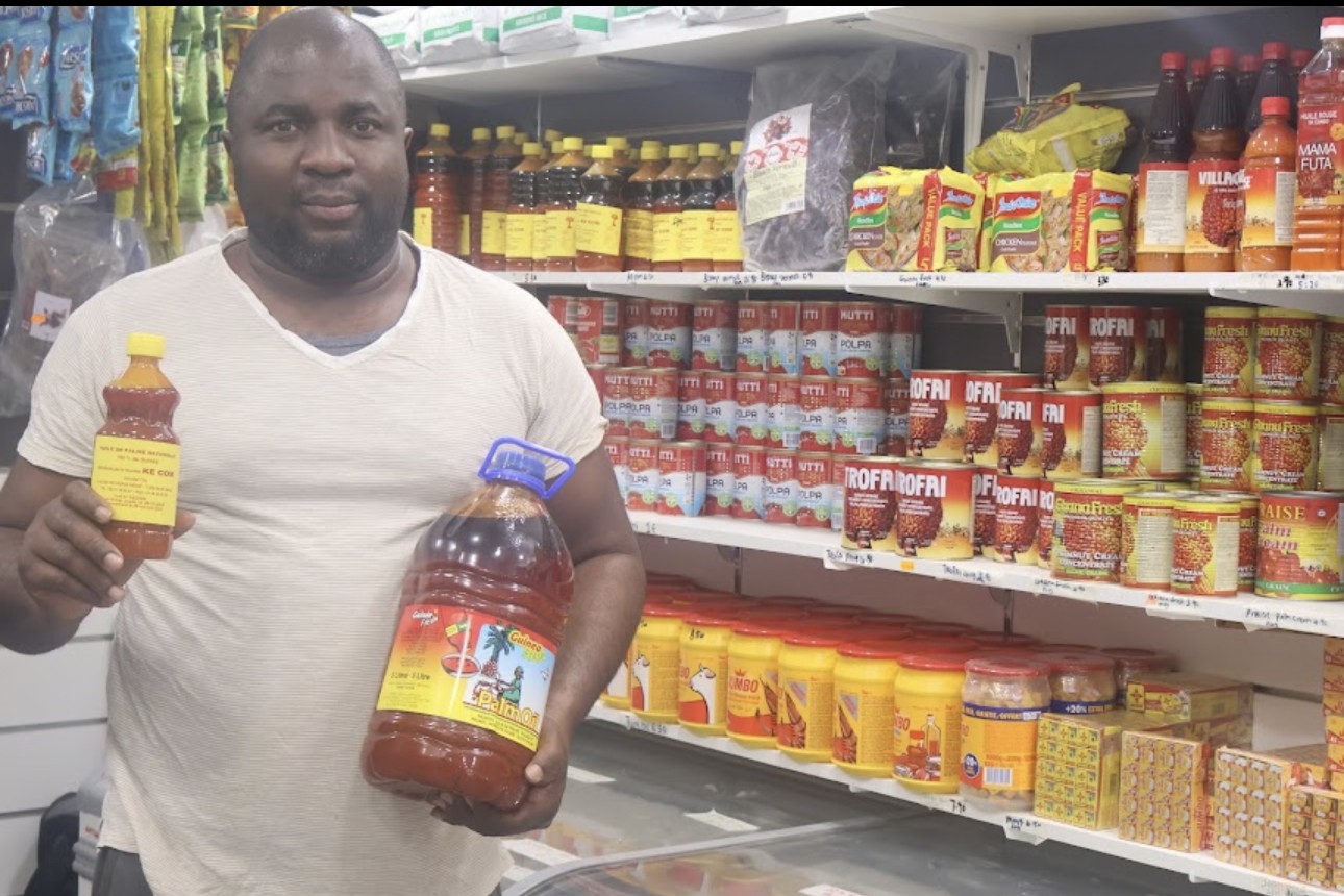 TEGA AFRO STORE shopkeeper holding palm oil in front of stocked grocery shelves
